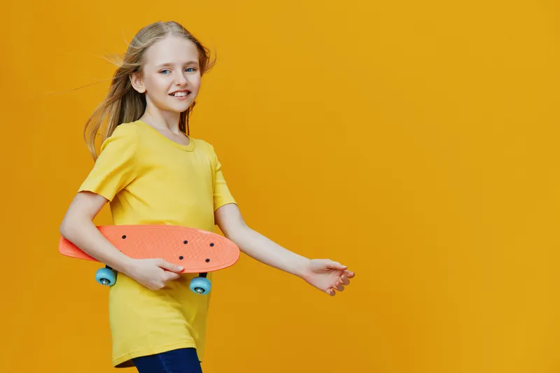 Young girl with long hair in a yellow t shirt holding an orange skateboard, smiling against a vibrant yellow background, embodying youth and freedom