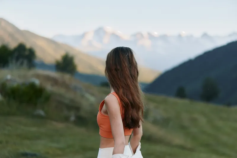 A woman with long hair wearing an orange top stands in a scenic mountain meadow, gazing toward distant peaks. The serene landscape radiates calm and natural beauty.