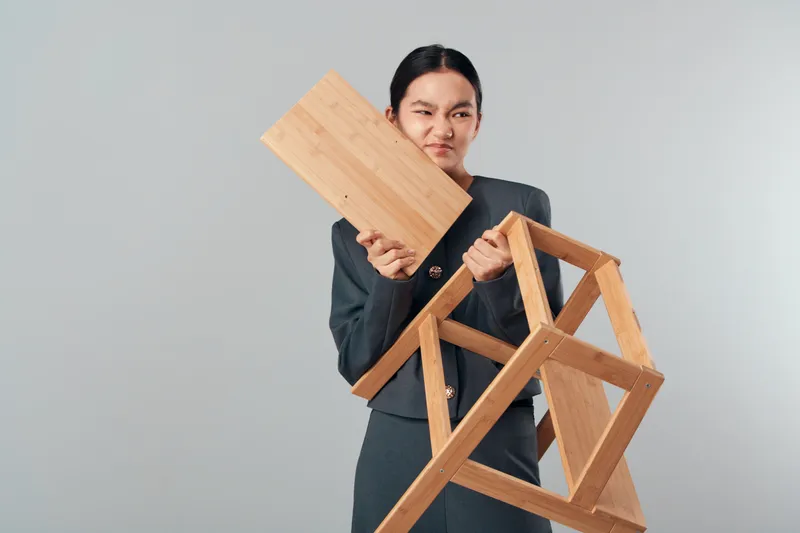 Young woman in casual outfit holding broken wooden chair and plank with frustrated facial expression isolated on gray background. Repair idea and furniture damage concept in studio.