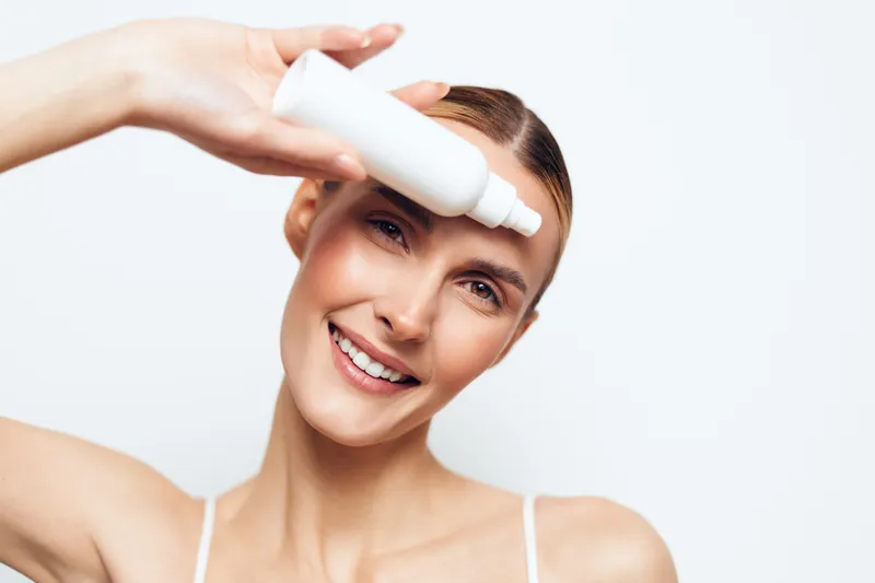 Young woman applying eye drops, smiling with clear skin, natural makeup, white background, closeup portrait, healthcare concept, fresh and clean appearance.