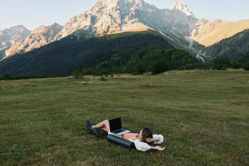A relaxed freelancer lies on a grassy meadow with a laptop in a rugged mountain landscape. The person seems focused and content, enjoying remote work amid nature and fresh air.