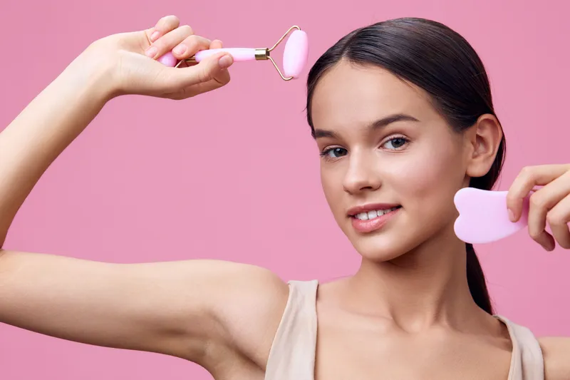 Young woman with long hair joyfully using a pink facial roller and heart shaped tool on a pastel pink background, showcasing beauty and skin care