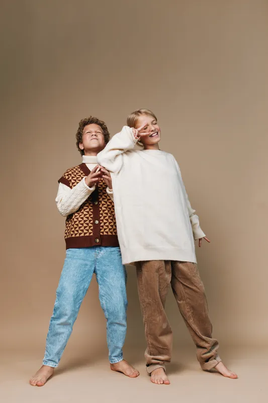 Two friends pose in cozy sweaters on a neutral backdrop, showcasing casual fashion, warm textures, relaxed joy, barefoot stance, and smiles captured in soft studio lighting.