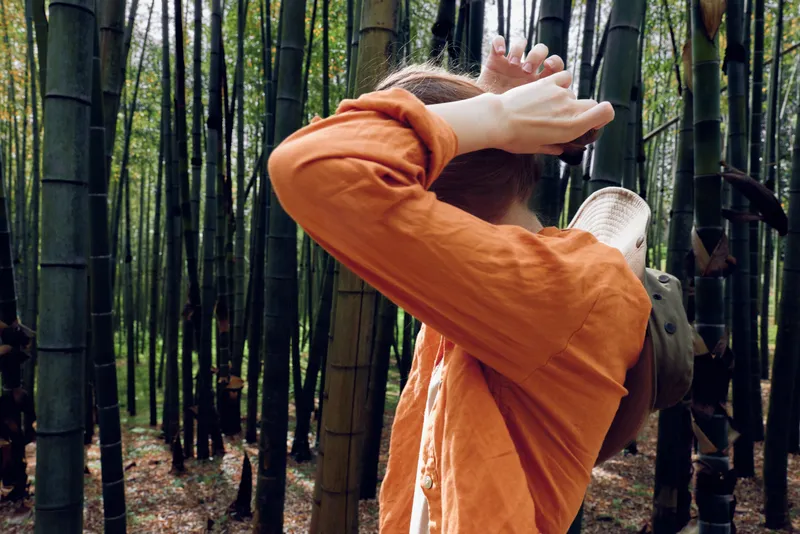 Bamboo forest person in an orange shirt with a hat resting on the back of their neck, standing among tall bamboo trunks in a contemplative pose during a nature walk outdoors.