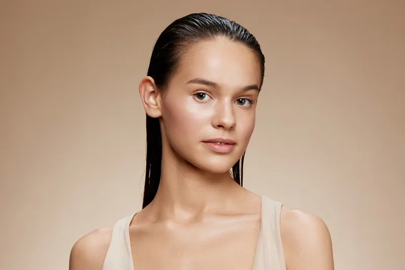 Natural beauty portrait of a young woman with wet hair, showcasing a fresh, glowing complexion against a beige background, ideal for skincare and beauty concepts