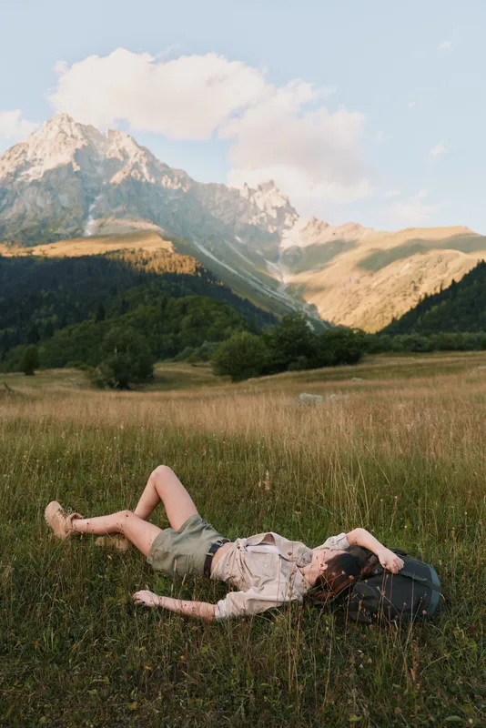 Young woman with long hair lies relaxed on grass in a mountain meadow wearing casual clothes and a hat enjoying nature and tranquility on a sunny day.