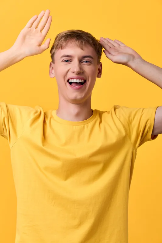Joyful, cheerful man in a yellow shirt raises hands in a bright studio, exuding happiness and energy, a celebration vibe with a smiling expression against a vivid background