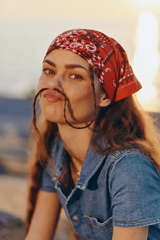 Lifestyle woman with modern boho-western denim street style wearing red bandana, playful expression, warm film color tones, outdoor natural light, casual summer vibe.
