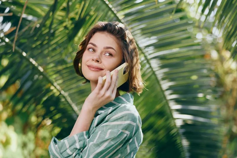 Young woman outdoors talking on a cellphone, smiling softly while surrounded by lush palm leaves and bright natural light.