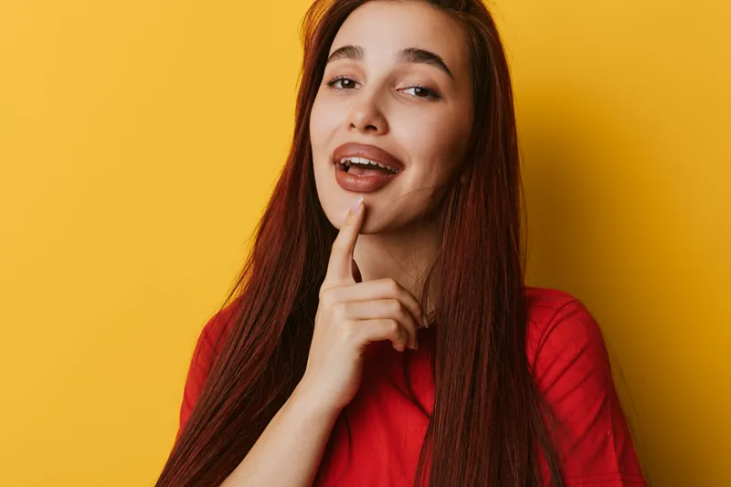 Young woman with long hair and braces smiling and touching chin on yellow background. Portrait of happy girl in red shirt with confident expression and casual pose.