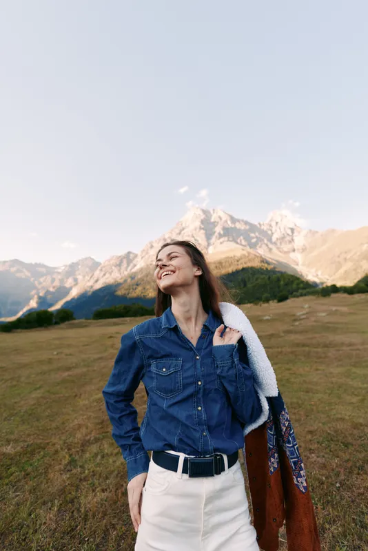 Woman portrait outdoors in a meadow with mountains behind, with a bright smile, wearing a denim shirt and carrying a jacket over shoulder, casual fashion pose capturing joyful nature and freedom.