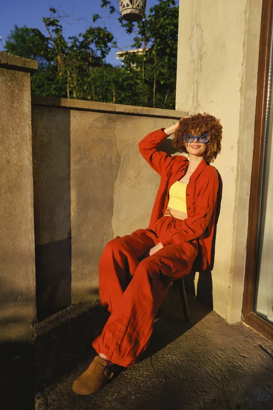 Woman lifestyle shot of a relaxed woman sitting on a balcony in a red suit and yellow top, sunny outdoor setting, casual chic pose, stylish afternoon mood
