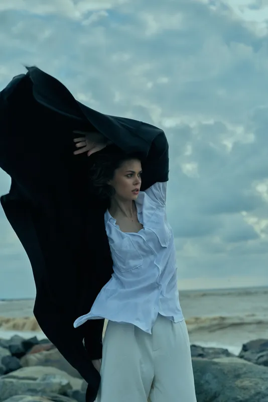 Woman on a windy beach lifts a dark cape over a white blouse, dramatic motion against a cloudy sea scene, fashion mood and movement