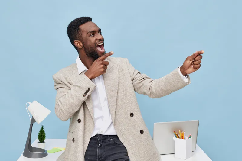 Confident cheerful young black man in beige blazer pointing and smiling while talking in modern office with laptop and desk accessories, capturing a lively and positive work environment. People lifest