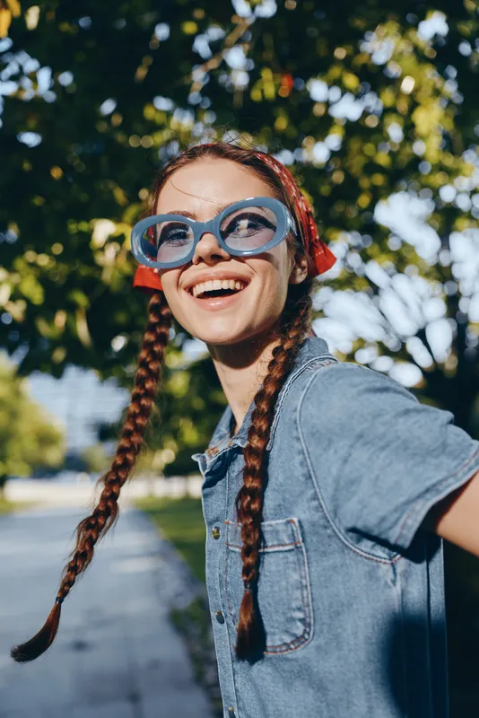 Lifestyle woman in modern boho-western denim street style wearing blue oversized glasses, smiling joyfully with braided hair under natural sunlight, film color effect.