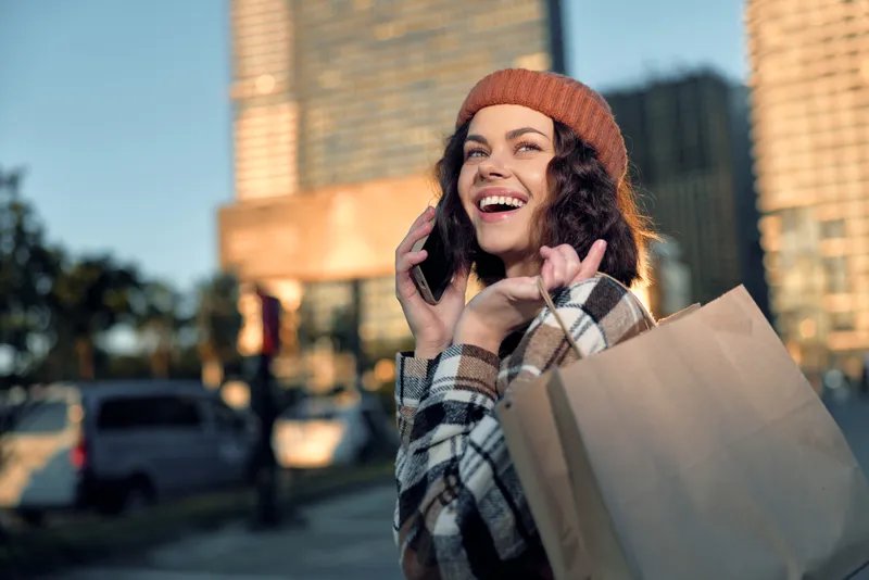 Woman shopping with phone and smile in urban street holding bags during golden hour glow, candid lifestyle moment with authenticity, mindful living and emotional storytelling in warm city light.