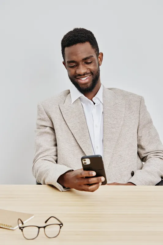 Smiling man using mobile phone, casual beige blazer, white shirt, young African-American male, happy emotions, modern office background, indoor shot, lifestyle concept, blurred neutral backdrop.