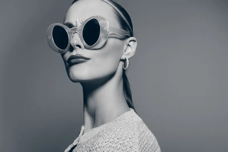 Stylish white woman with pale skin and slicked back hair wears large round sunglasses and a textured sweater. Portrait in monochrome with confident expression and studio lighting.
