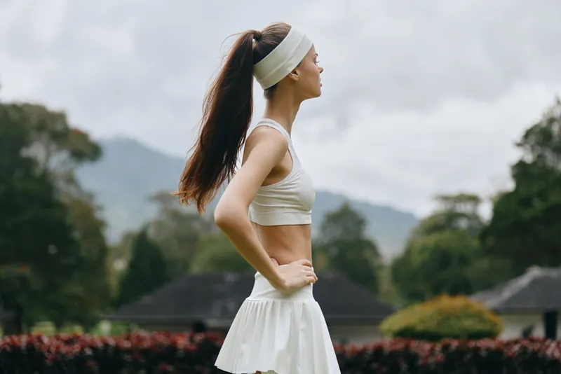 Young athletic woman in white sportswear posing outdoors, expressing confidence, with a serene mountain background, perfect for fitness and wellness concepts
