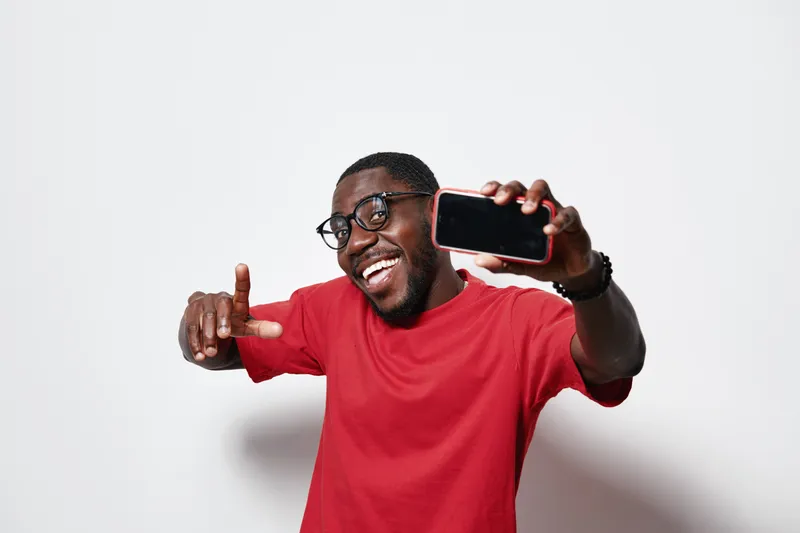 Black man with dark skin wearing glasses and red shirt smiling and holding smartphone taking selfie on white background. Happy African male posing with phone for photo.