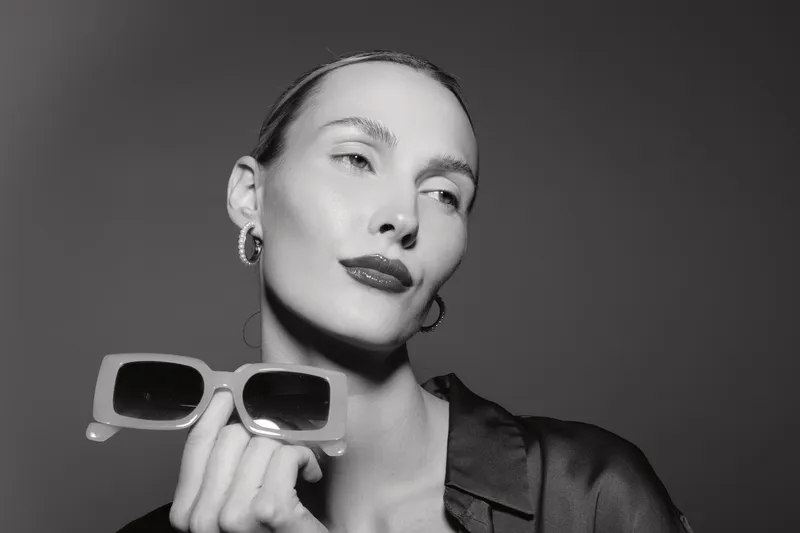 Caucasian woman with pale skin holds sunglasses, posing confidently in black attire with simple earrings, studio portrait in monochrome.