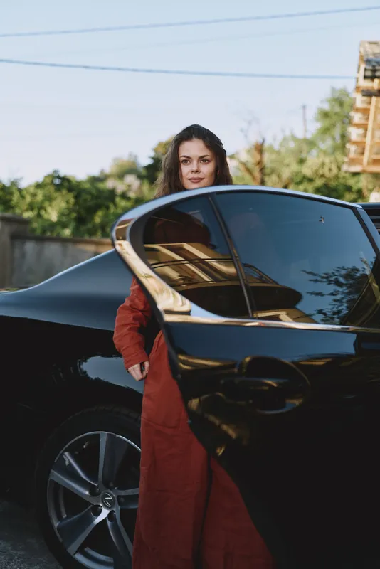 Woman in a red coat stands beside a sleek black car, posing with the open door, capturing a confident lifestyle moment outdoors and a modern urban mood