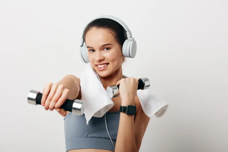 Young woman in sports bra and towel over shoulders holding dumbbells, wearing headphones and smartwatch, smiling during indoor fitness workout on clean white background, active sport concept.