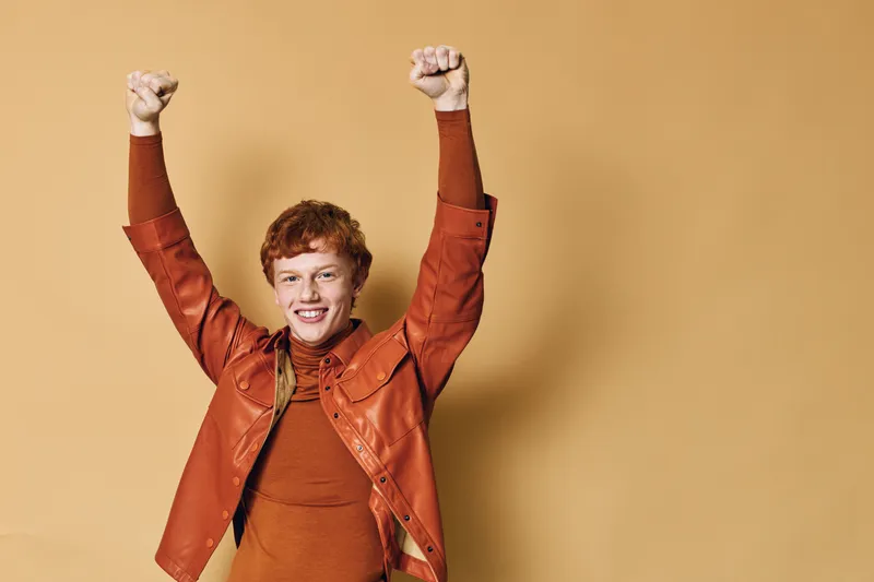 Redhead woman with curly hair wearing orange jacket and sweater, raising fists celebrating success or achievement with smile on beige background studio portrait.