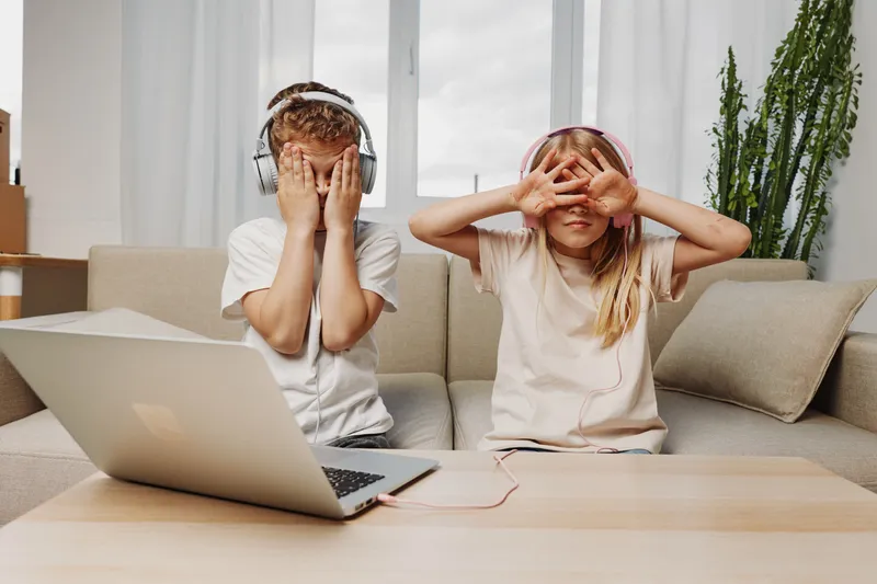Two children wearing headphones playfully cover their faces sitting on a couch with a laptop on the table in bright living room environment.
