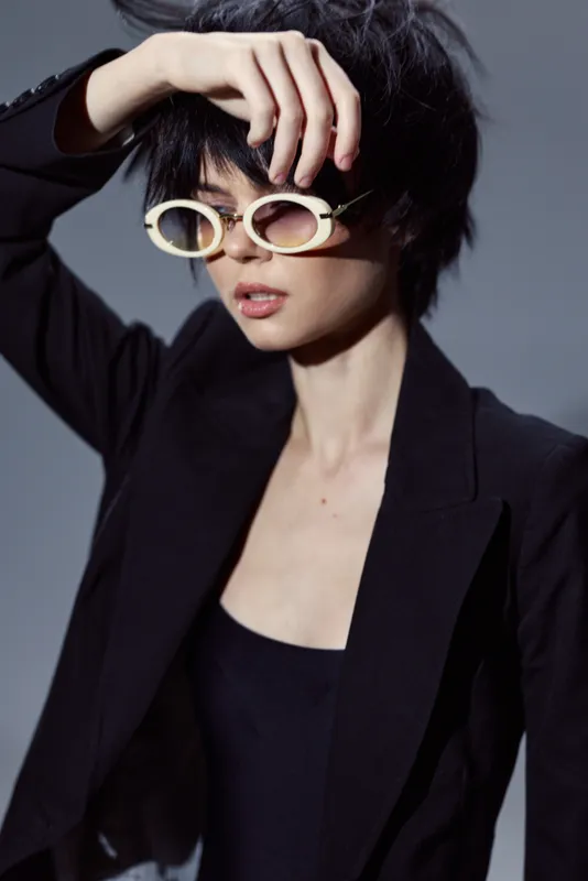 Stylish woman wearing white sunglasses and black blazer posing with hand on forehead