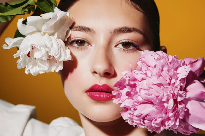 Close-up of a young woman with bright red lips holding white and pink flowers near her face. Bold makeup and creative beauty styling on a colorful studio background, fashion concept.