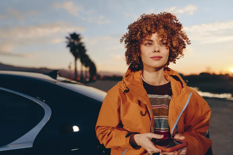 Woman lifestyle outdoors, curly hair, orange jacket, standing beside car at sunset evening, holding smartphone, warm golden light, calm confident expression, coastal road background