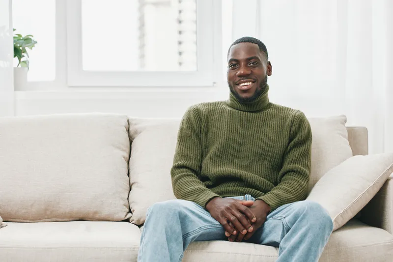 Smiling Black man in a cozy green turtleneck sweater sitting on a neutral sofa in a bright, airy room, conveying a sense of comfort and relaxation
