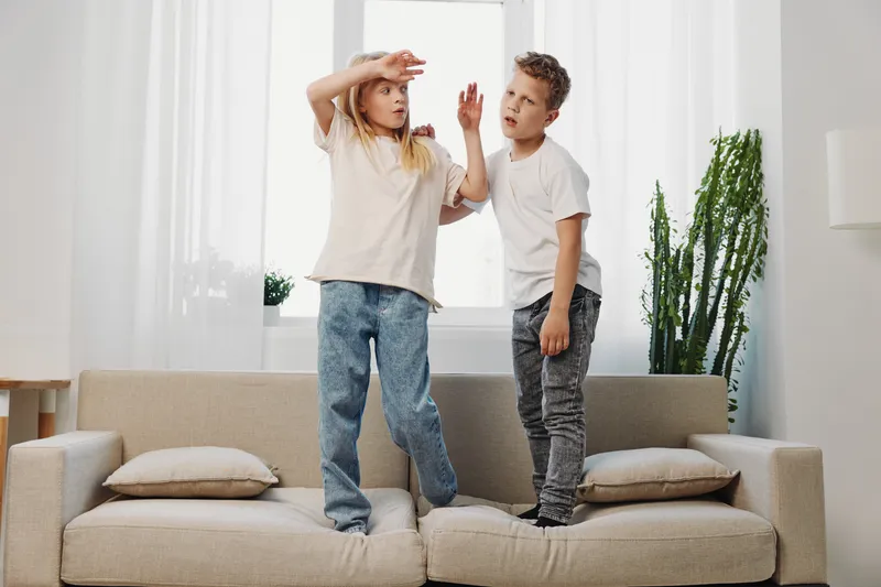 Two kids standing on a couch indoors, showing playful expressions and gestures in a bright living room with cushions and plants. Concept of childhood fun and home activity.
