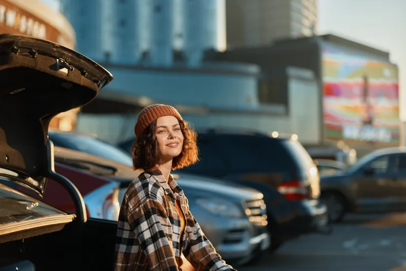 Woman by car trunk in urban parking, smiling in a candid lifestyle portrait at golden hour glow with beanie and plaid shirt, authenticity, mindful living and emotional storytelling.