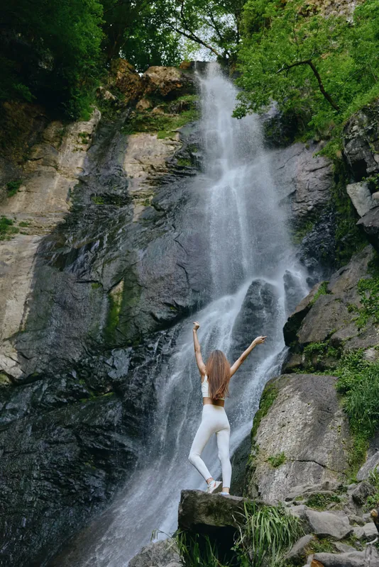 woman waterfall enjoying nature, wearing white activewear and standing on rock surrounded by green forest and cascading water, outdoor adventure and freedom