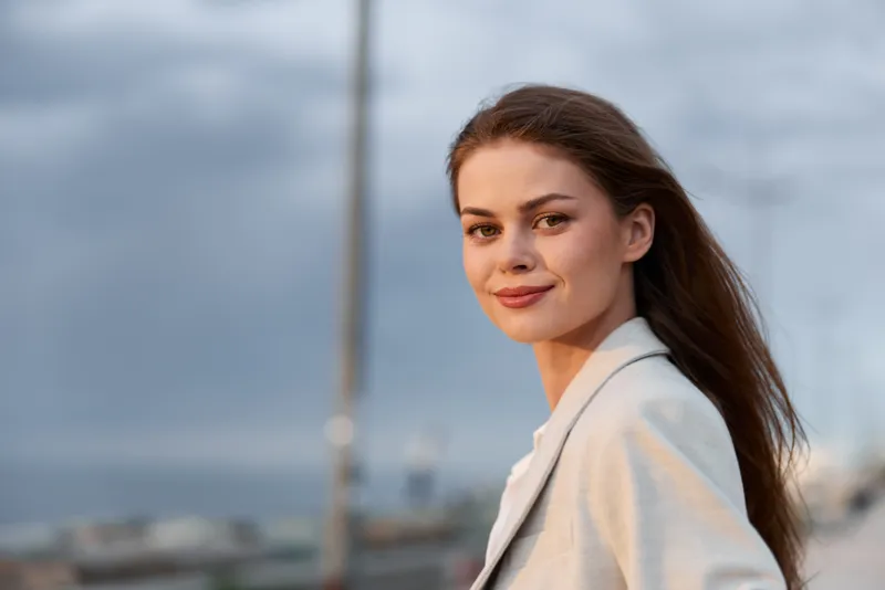 woman confident smiling portrait with long hair in light jacket on blurred urban background during golden hour lifestyle fashion