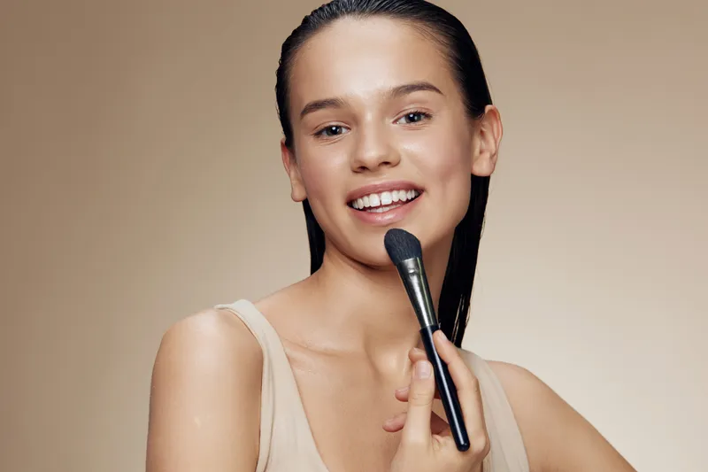 Young woman applying makeup with a brush, showcasing natural beauty and radiant skin, ideal for beauty and skincare concepts