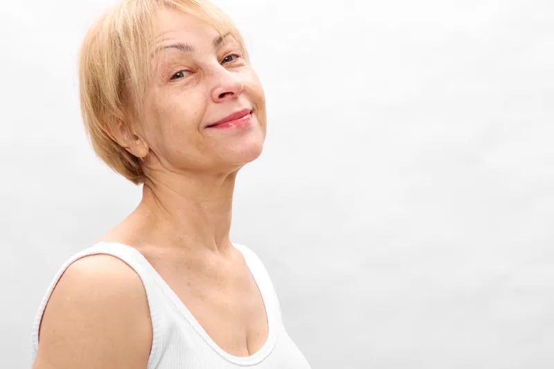 Senior woman, smile portrait, side view, short blonde hair, relaxed posture, soft daylight, indoor white background, calm expression, wellbeing