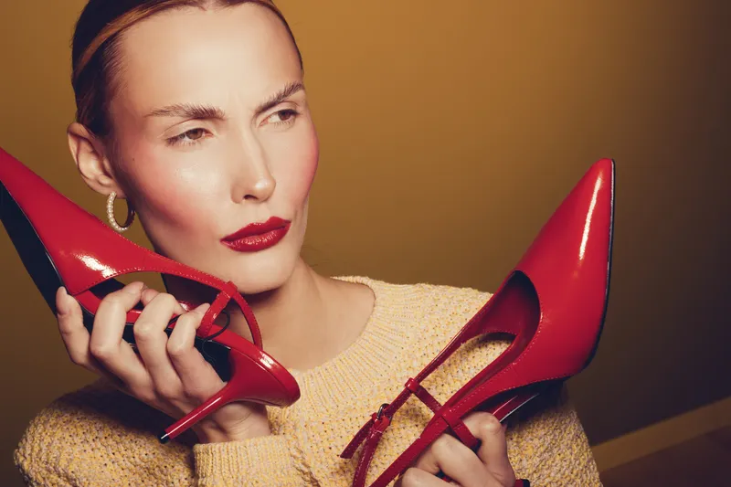 Woman with red lipstick holds red high heel shoes near face, wearing beige sweater and gold hoop earrings on brown background. Concept of fashion, style, and accessories.