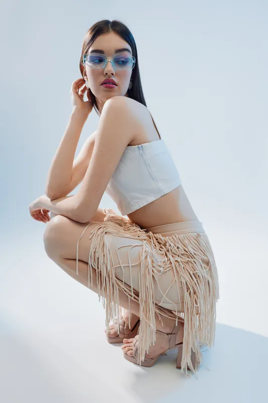 Young woman in stylish sunglasses and a white crop top poses while crouching. She wears fringed pants and heels in a bright studio. Concept of woman beauty fashion and modern style.