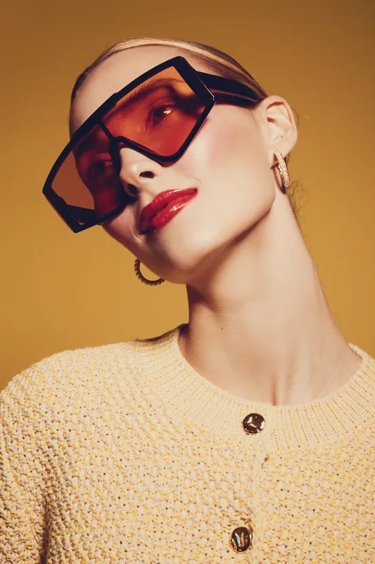 Stylish woman with short blonde hair wears large tinted sunglasses and red lipstick. She poses confidently against a warm yellow background in a textured sweater. Fashion and beauty concept.