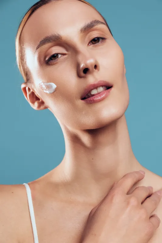 A close-up portrait of a young woman with clear skin and a small amount of cream on her cheek poses against a blue background, showcasing skincare and natural beauty concepts.