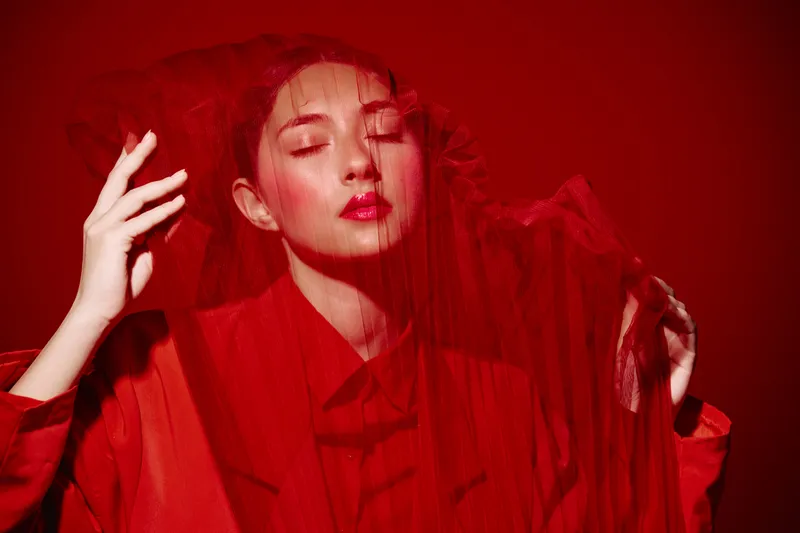 Young woman with closed eyes posing behind translucent red fabric wearing a red coat liught softly on a red background. Concept of beauty fashion creative elegance.