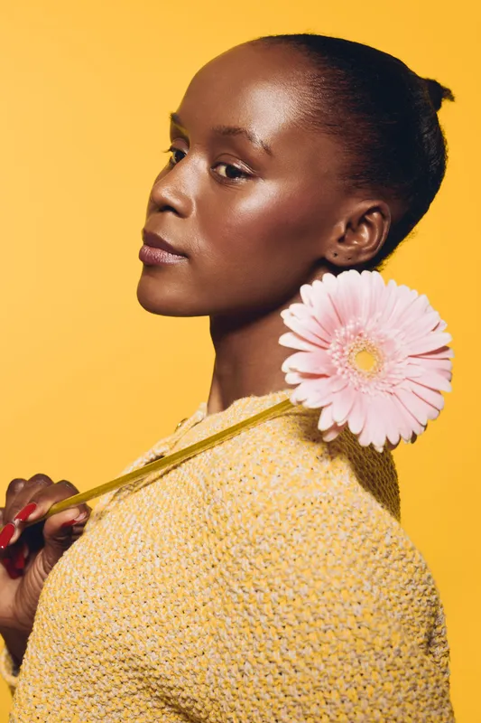 Portrait of a confident dark skinned African woman holding a pink flower over her shoulder. Yellow background and knitted sweater create a warm, stylish concept of beauty and fashion.
