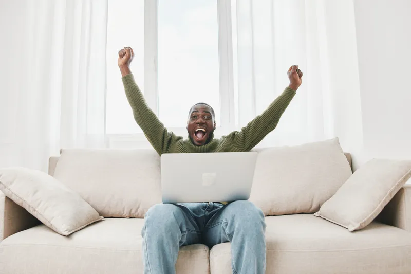 Excited African man celebrating success at home, raising arms in joy while sitting on a sofa with a laptop Cozy interior with soft colors, capturing a moment of triumph