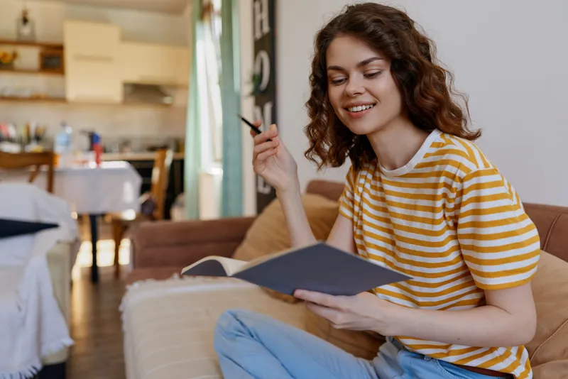 A smiling woman sits on a sofa at home, reading a notebook and holding a pen. A cozy living room scene conveying study, creativity, and personal organization in a warm, relaxed atmosphere.