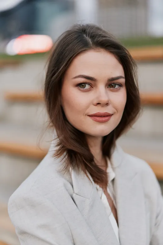 confident woman in beige blazer with subtle smile sitting outdoors with blurred urban background, natural light, neutral tones, fashion concept