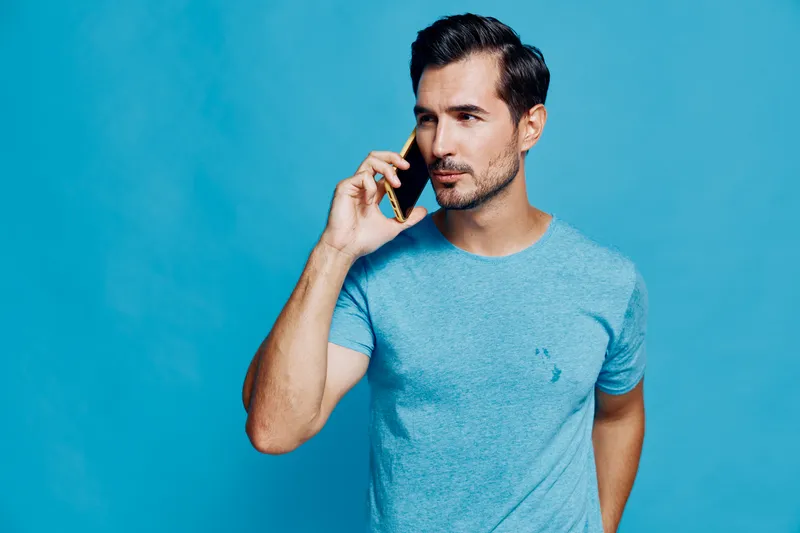 Handsome man talking on mobile phone while standing against a bright blue background. Confident, stylish, and focused on conversation in casual attire.