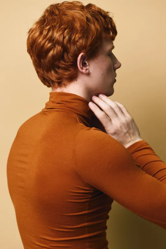 Redhead young man in rust turtleneck sweater posing sideways with hand near neck on beige background. Profile portrait of fashionable male with curly hair and casual style indoors.